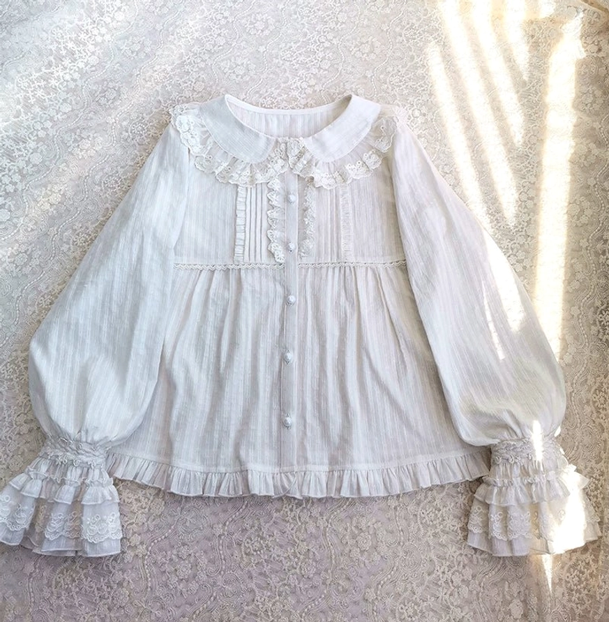 White long-sleeve lolita cotton blouse with peter pan lace collar, tiered ruffle cuffs, and pleated front detailing, displayed on lace fabric with sunlight shadows, showcasing relaxed a-line silhouette and vertical striped cotton texture