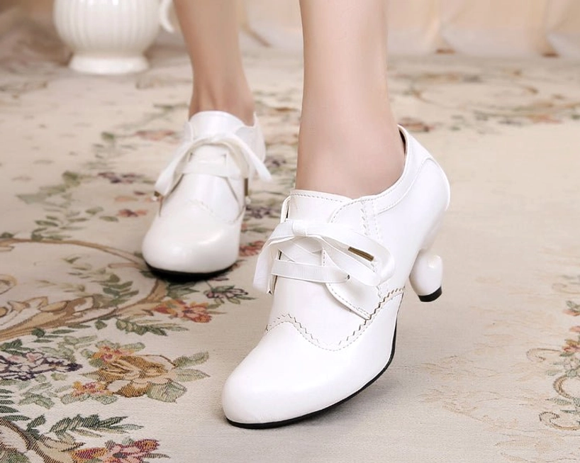 Pure white retro lace-up Lolita high heel shoes with solid white finish, scalloped stitching trim, curved spool heel, and ribbon lace fastenings, worn on indoor floral patterned carpet ground view