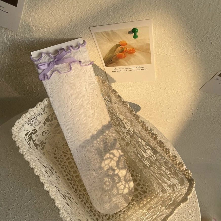 White ruffled lace ankle socks with purple ribbon bow accent worn on model's leg showing delicate lace texture and sweet ruffled sock cuff trim