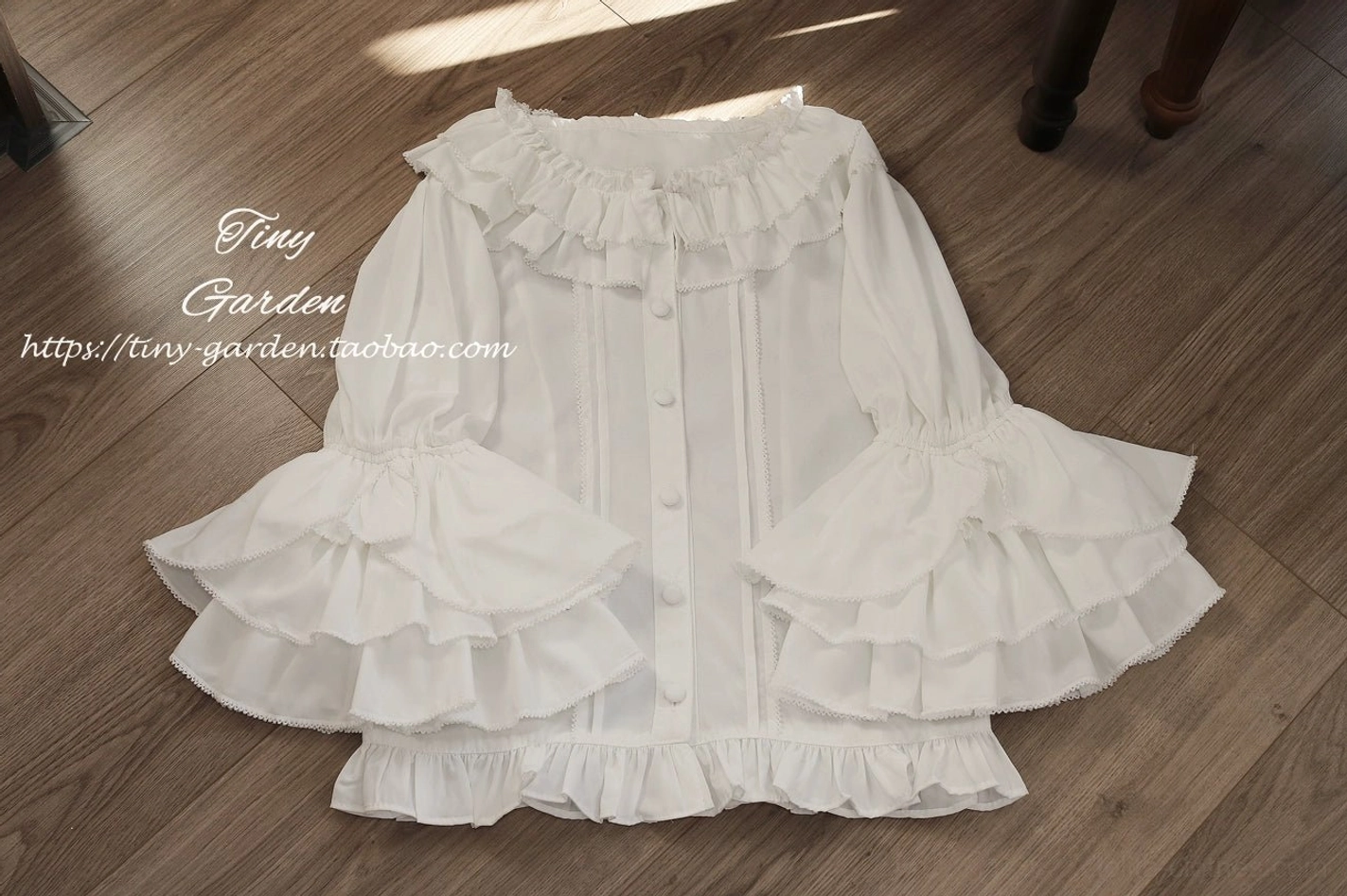White vintage ruffled lolita blouse in flat lay top-down view on wooden table background, showing ruffled collar, layered bell sleeves, front button details and ruffled hem construction