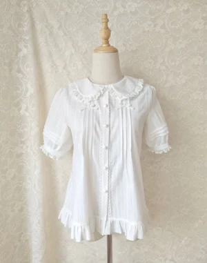 White short-sleeve sweet lolita blouse with ruffled peter pan collar, front button placket, chest bows, and ruffled cuffs, displayed on mannequin with lace backdrop