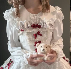 Close-up front upper body view of white sweet Lolita blouse showing lace ruffled Peter Pan collar with red bow front decorations and white satin fabric details