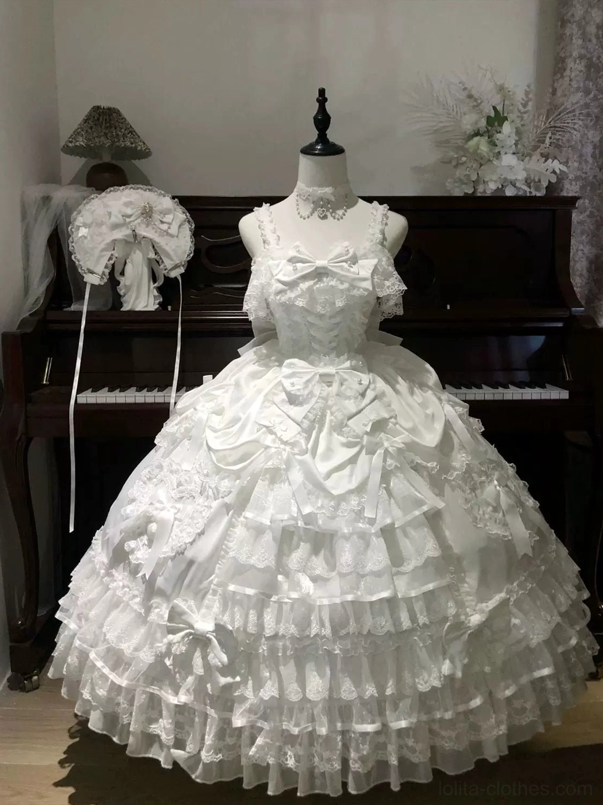 White sweet wedding Lolita JSK dress on dress form indoor vintage piano studio setting with high neck lace trim, satin bow on bodice, full multi-layered ruffle lace skirt, white bridal headdress displayed beside
