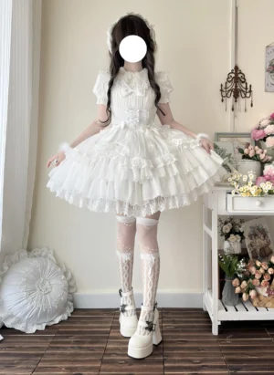 White sweet lolita JSK dress with tiered ruffled hem and lace trim, worn by model in vintage flower shop setting, showcasing peter pan collar and short puff sleeves front view