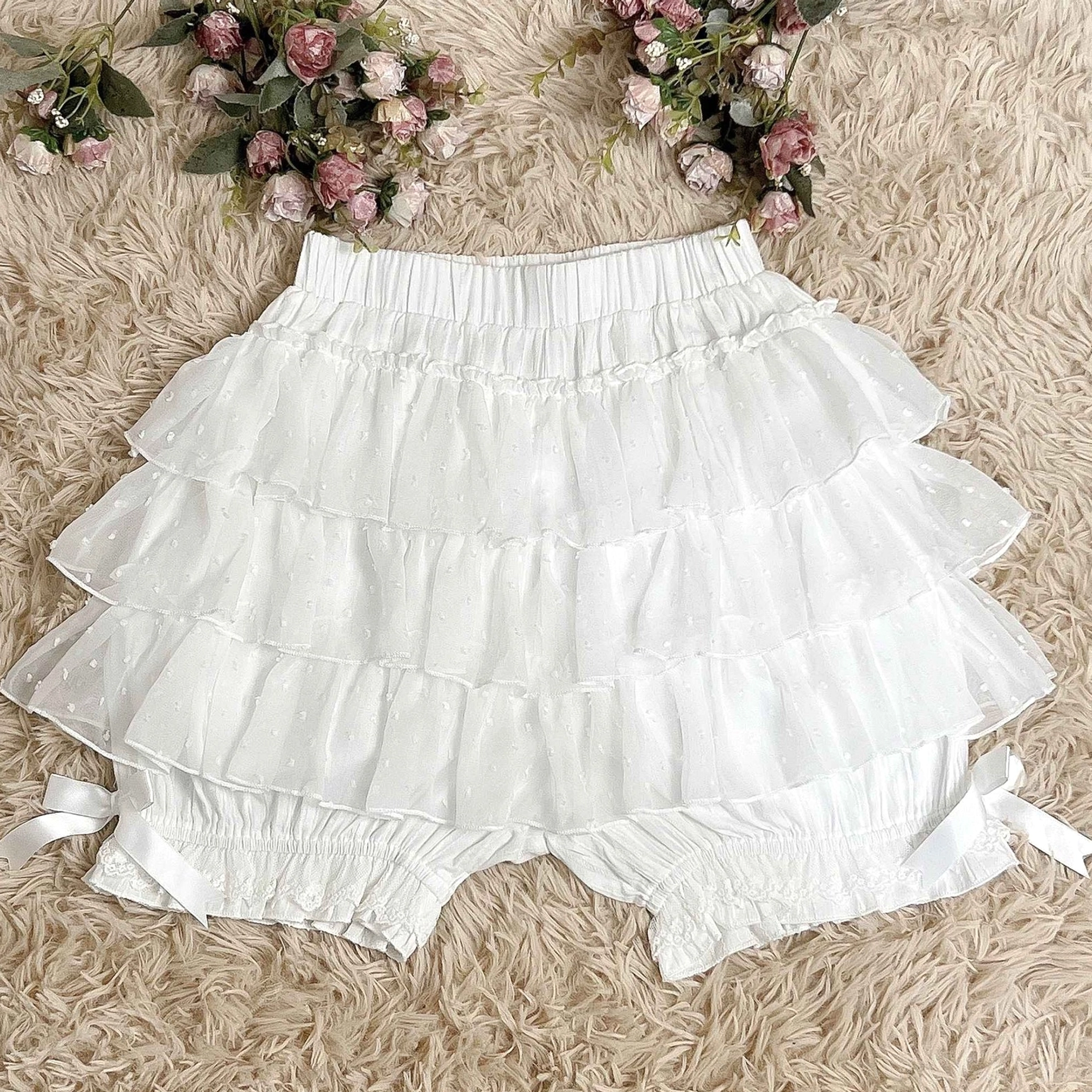 White tiered ruffle lolita bloomers overhead flat lay view on furry fabric background with artificial flower decorative props showing layered ruffles lace trims and side ribbon details