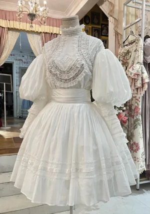 White vintage Victorian Lolita two-piece set with ruffled high stand collar, handmade lace embroidery on bust, oversized puff sleeves, and pleated skirt displayed on dress form in boutique setting