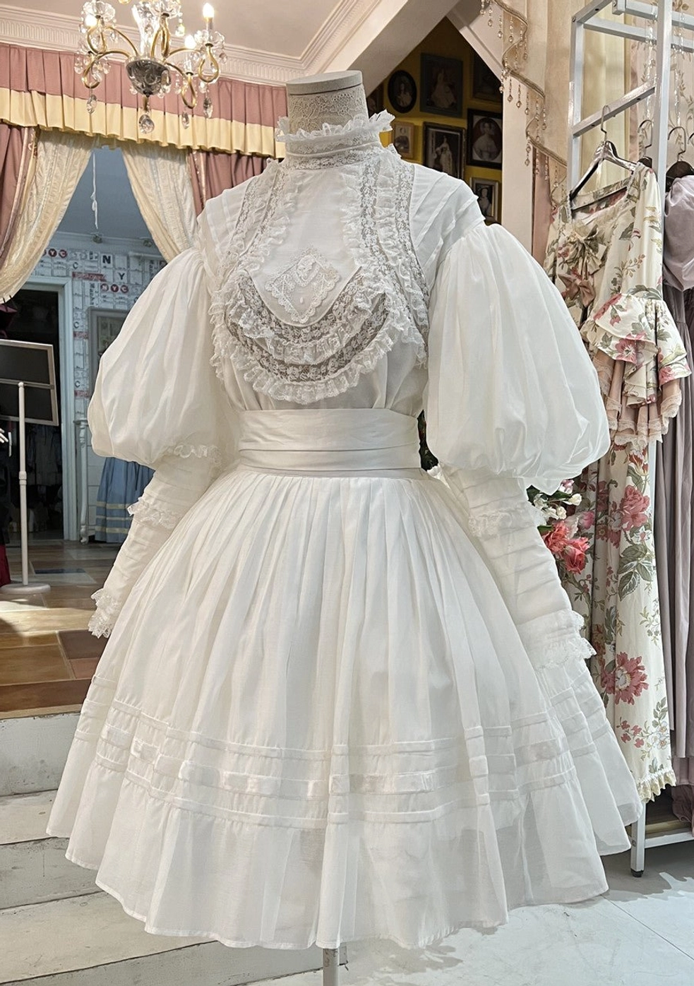 White vintage Victorian Lolita two-piece set with ruffled high stand collar, handmade lace embroidery on bust, oversized puff sleeves, and pleated skirt displayed on dress form in boutique setting