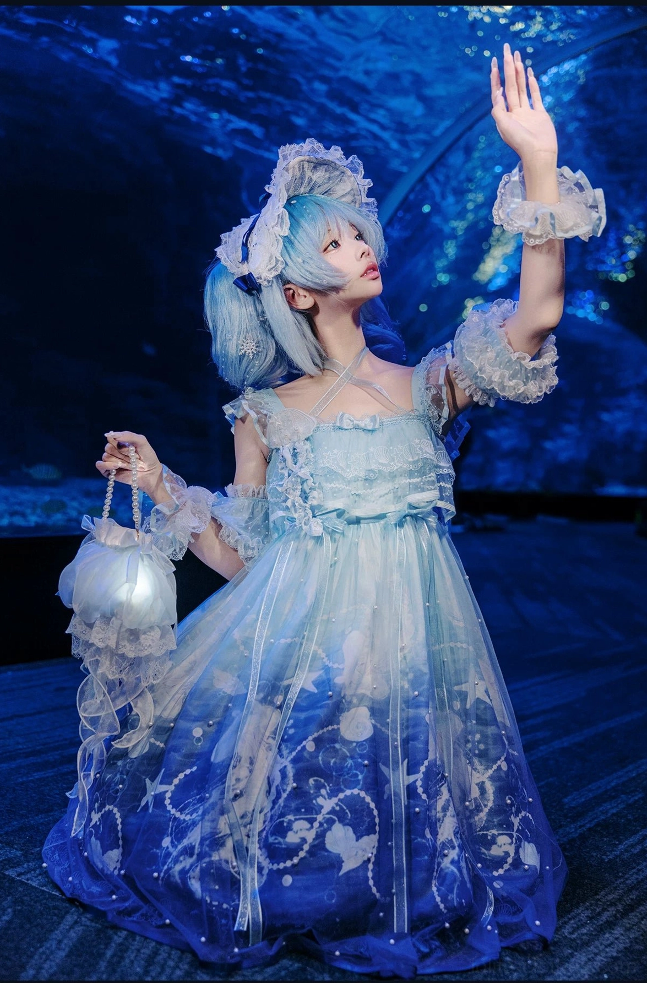 Three-quarter side profile shot of model in blue gradient jellyfish-themed sweet lolita complete outfit with ocean embroidery, matching headdress and handbag in underwater aquarium setting