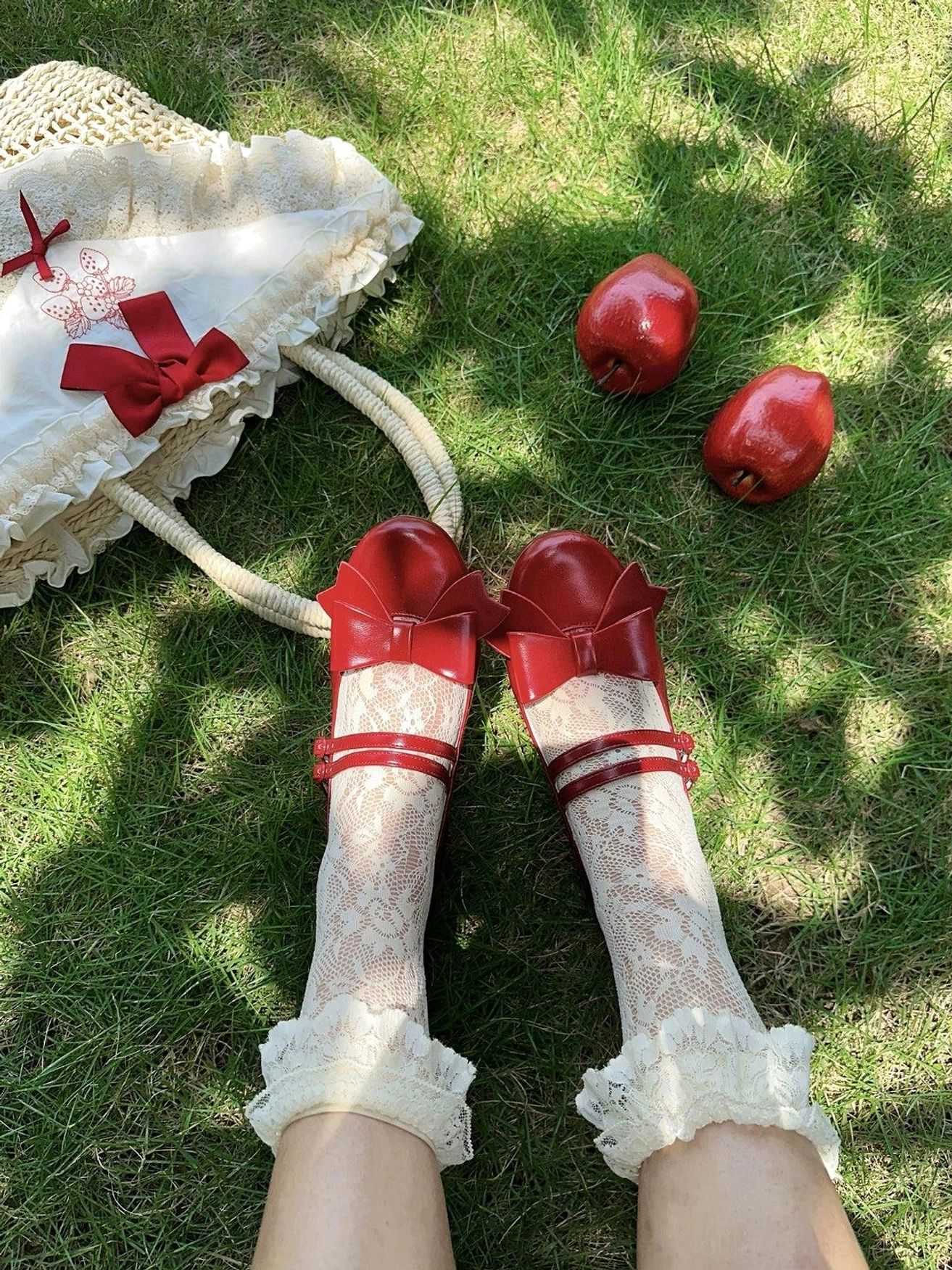 Wine red Lolita Mary Jane shoes worn on feet first-person overhead view on sunlit outdoor lawn with ruffled lace socks strawberry props and straw handbag styling