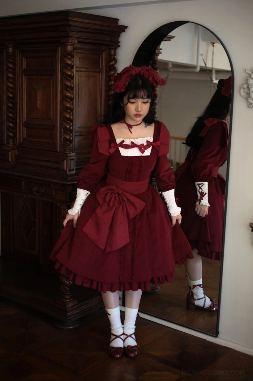 Burgundy wine red velvet Lolita OP dress mirror side profile full body shot in vintage wooden furniture setting, displaying matching red hair bow accessory and dress silhouette from side perspective