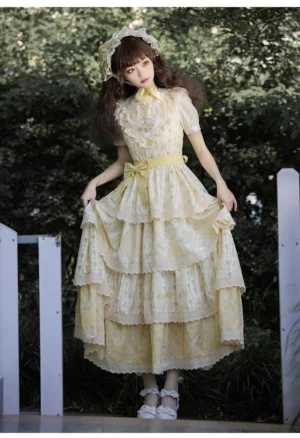 Full-body front view of yellow cream embroidered country Lolita JSK dress with short puff sleeves, multi-tiered ruffled skirt featuring lace trims, and coordinating yellow waist bow, modeled outdoors in garden setting with white picket fence background