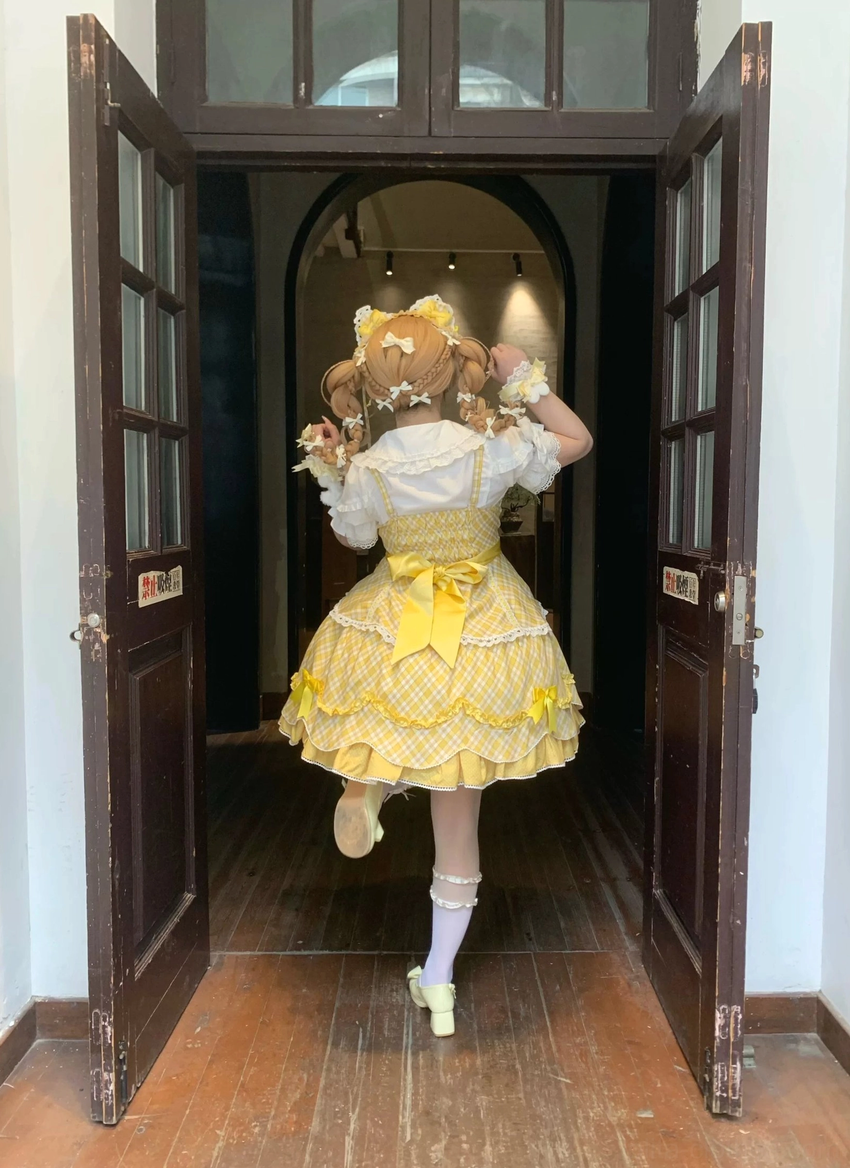 Yellow plaid sweet lolita OP one-piece dress back full-body view showing large back waist detachable bow, tiered ruffled skirt hem with lace accents, vintage wooden door hallway scene, model wearing bunny ear hair accessory and light green flat shoes